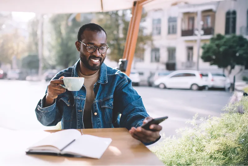 Smiling man sitting, drinking coffee at a table while looking at his phone