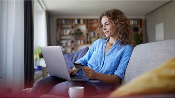 Woman at home beside a cup of coffee entering account information on a laptop from a bank card she is holding